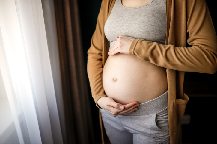 Unrecognisable woman embraces her pregnant belly by the window in soft moody light.